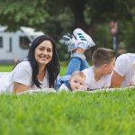 Happy family lying on grass with RV in the background, enjoying a sunny outdoor picnic.