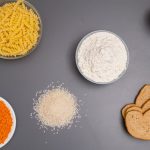 An assortment of dry ingredients including pasta, rice, and bread displayed on a grey background.
